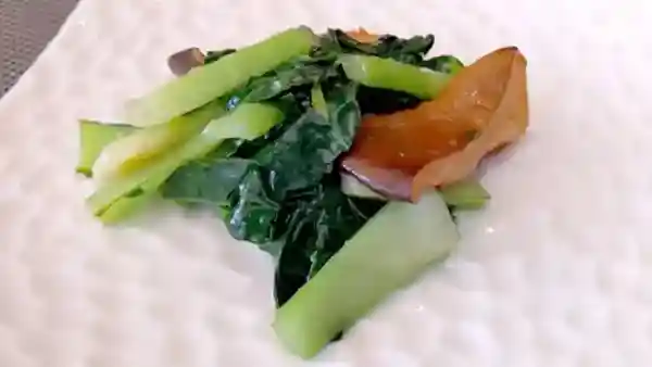 This photograph depicts stir-fried greens served on a white plate filled with brightly colored vegetables and brown porcini mushrooms.