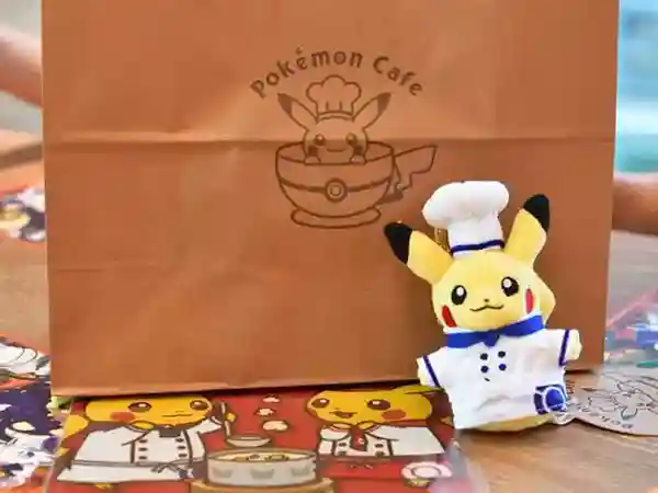 This photograph displays a paper bag containing exclusive Pokémon merchandise only available at the Pokémon Café. The bag is brown, measures 20 cm in height, and is 30 cm wide. The Pokémon Café logo is printed on the bag's surface, featuring an emblem of Pikachu dressed in his chef's outfit.