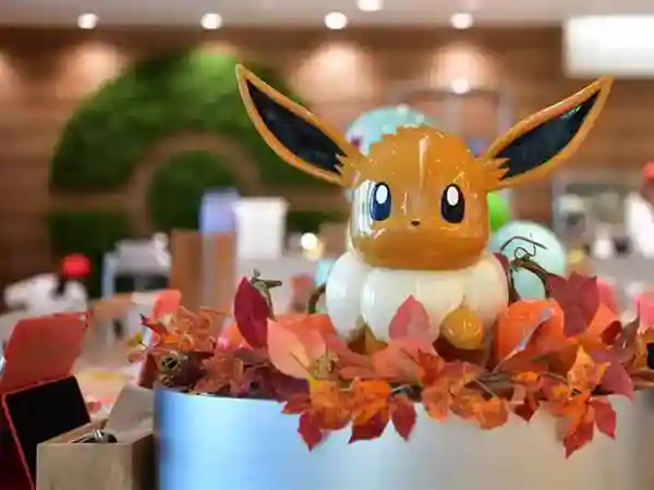 This photo depicts an Eevee doll that stands about 50 cm tall. The doll is displayed inside the Pokemon Cafe, surrounded by autumn leaves at its feet.