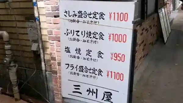 The picture displays a sign for Sanshuya Ginza located on Namiki-dori Avenue in Ginza 2 Chome. The poster measures approximately 1 meter in height and 80 centimeters in width. The prices for four set menus are written in red on a whiteboard. The prices are as follows: assorted sashimi for 1,100 yen, yellowtail teriyaki for 1,000 yen, grilled fish for 950 yen, and various fried fish for 1,100 yen.