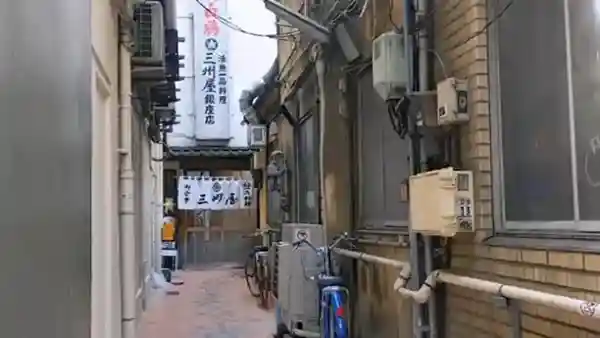 This photograph depicts the alley where the Sanshuuya Ginza store is located. The path has a width of 2 meters and a depth of 20 meters, with buildings lining both sides. At the end of the alley, you can see the entrance to the restaurant, marked by a white Noren curtain with "御食事 三州屋（Goshokuji Sanshuya）" written in Japanese.