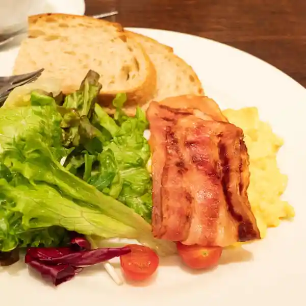 This photo shows a plate of the scrambled eggs set. It consists of scrambled eggs, grilled bacon, salad, and two slices of toast on a white plate. This toast is a bread called Minor Grain Life. This bread is baked with a blend of seven kinds of cereals.
