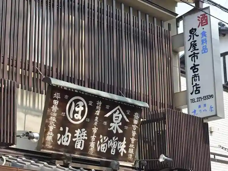 This photo shows the wooden sign for Izumiya Ichiko Shoten. The store's name is written in white letters on a brown board. In addition to the store name, soy sauce and miso are also documented.