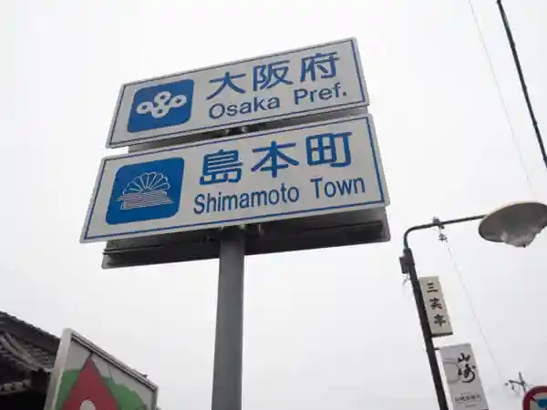 This photo shows a signboard on the border between Osaka and Kyoto prefectures. The words "Osaka Prefecture" and "Shimamoto Town" are printed in blue letters on white metal.