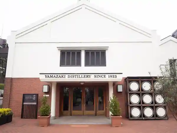 This photo shows the Whiskey Museum of Suntory Yamazaki Distillery. It is a white-walled building with whiskey barrels stacked by the entrance.