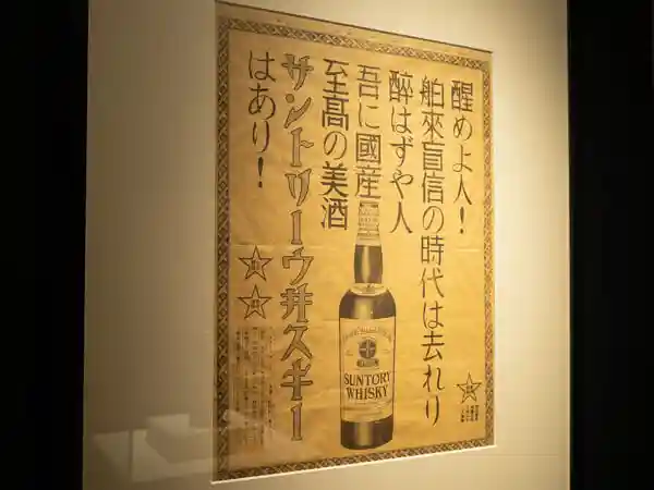 This photo is a poster of the first domestic whiskey poster of "Suntory White Tag." on display in the Whiskey Museum at Suntory's Yamazaki Distillery." It reads, "Wake up, people! The age of blind faith in imported spirits is over, and I have Suntory Whiskey, the supreme domestic sake!"