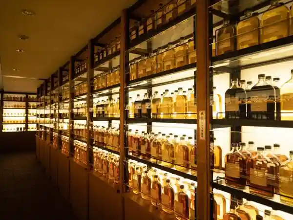 This photo shows the Whiskey Library in the Whiskey Museum at Suntory Yamazaki Distillery. Whiskey bottles are lined up on the wall. The bottles are lit up and shine in amber.