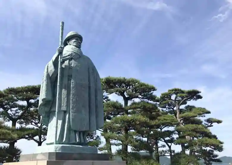 This photo shows a bronze statue of Kokichi Mikimoto at Mikimoto Pearl Island in Toba, Japan. The figure is depicted wearing a bowler hat and cloak and holding a cane in his right hand.