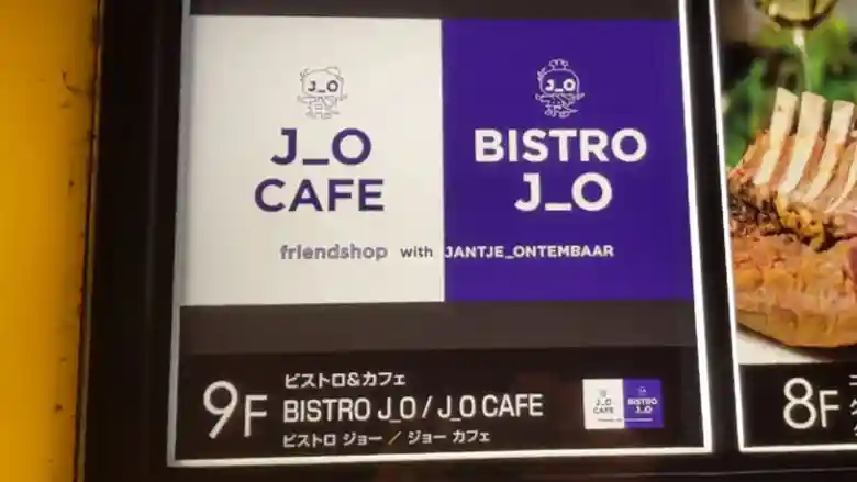 「銀座Velvia館」の入り口に掲げられた「BISTRO J_O」の看板です。紫と白色の背景にキャラクターが描かれています。