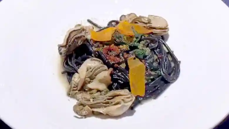 牡蠣のペペロンチーノ イカ墨を練り込んだスパゲティの写真です。丸く白い皿にイカスミが練り込まれた漆黒のパスタがもられています。表面には薄く切った黄色いカラスミが散らされています。
