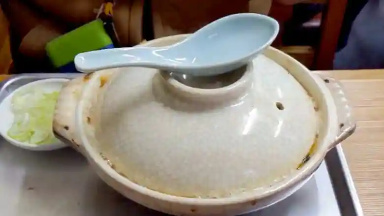 鍋焼きうどんの入った白色の土鍋の写真です。土鍋は蓋をした状態で、蓋の上に水色のレンゲが置かれています。