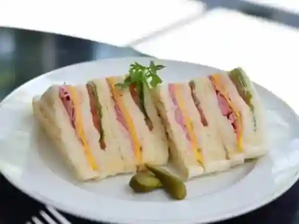 This photograph shows the American Clubhouse Sandwich served on a white plate on an outdoor table. The sandwich ingredients include smoked chicken, bacon, and egg, and the toasted bread has a charred surface.