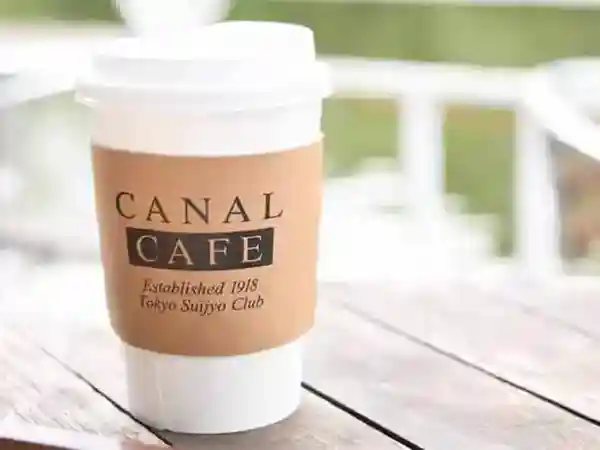 This photo displays a cup of coffee on a table on the Boat Deck, with the Ushigome moat in the background. The cup is white and has a light brown label with the words "CANAL CAFE" written in dark brown letters.