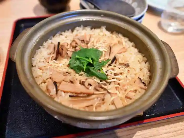 土鍋に入った松茸ご飯の写真です。お醤油に薄くそまったご飯の上に三つ葉と松茸が添えられています。