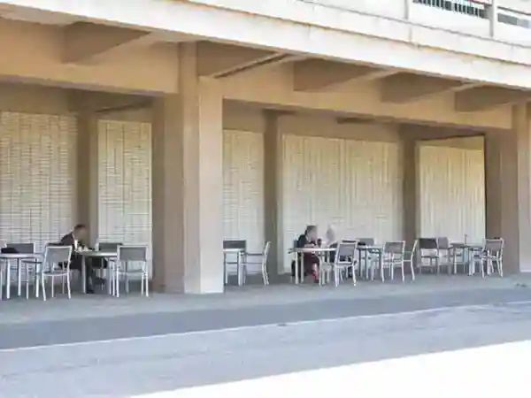 This photo shows tables and chairs in front of the Toyokan building. A few people can be seen working on their computers in the shade under the building's terrace.